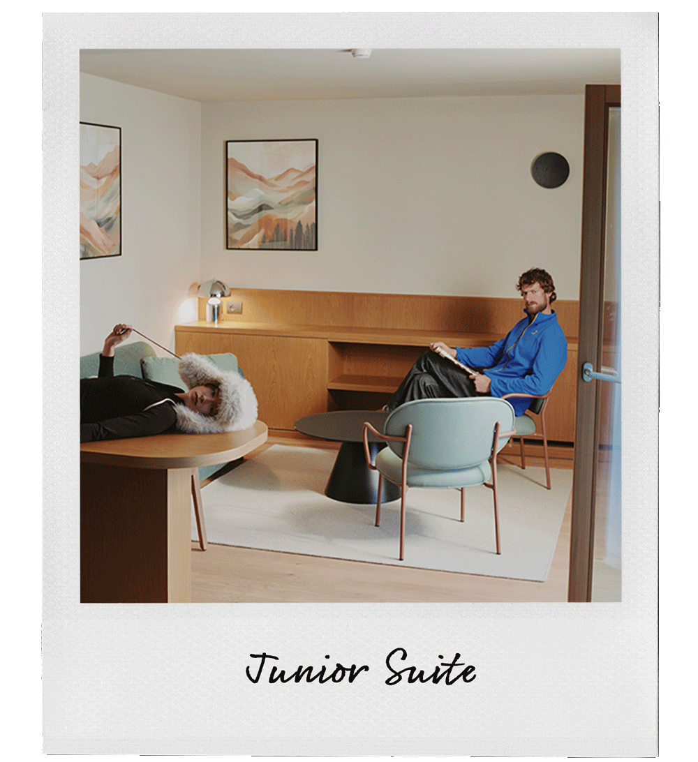 Interior of a hotel junior suite with two people, one lying on a couch and the other sitting in a chair, in a modern, minimalist decor with abstract landscape art on the walls.