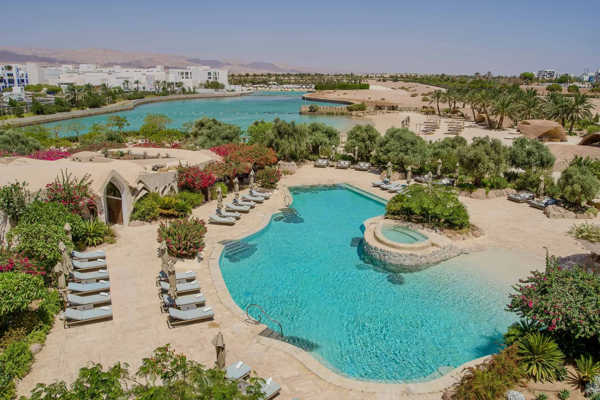 A luxurious resort with a large swimming pool surrounded by lounge chairs, lush green trees, and vibrant pink flowers. A calm lake or lagoon with a thin band of water separates the pool area from white buildings and desert mountains in the background under a clear blue sky.