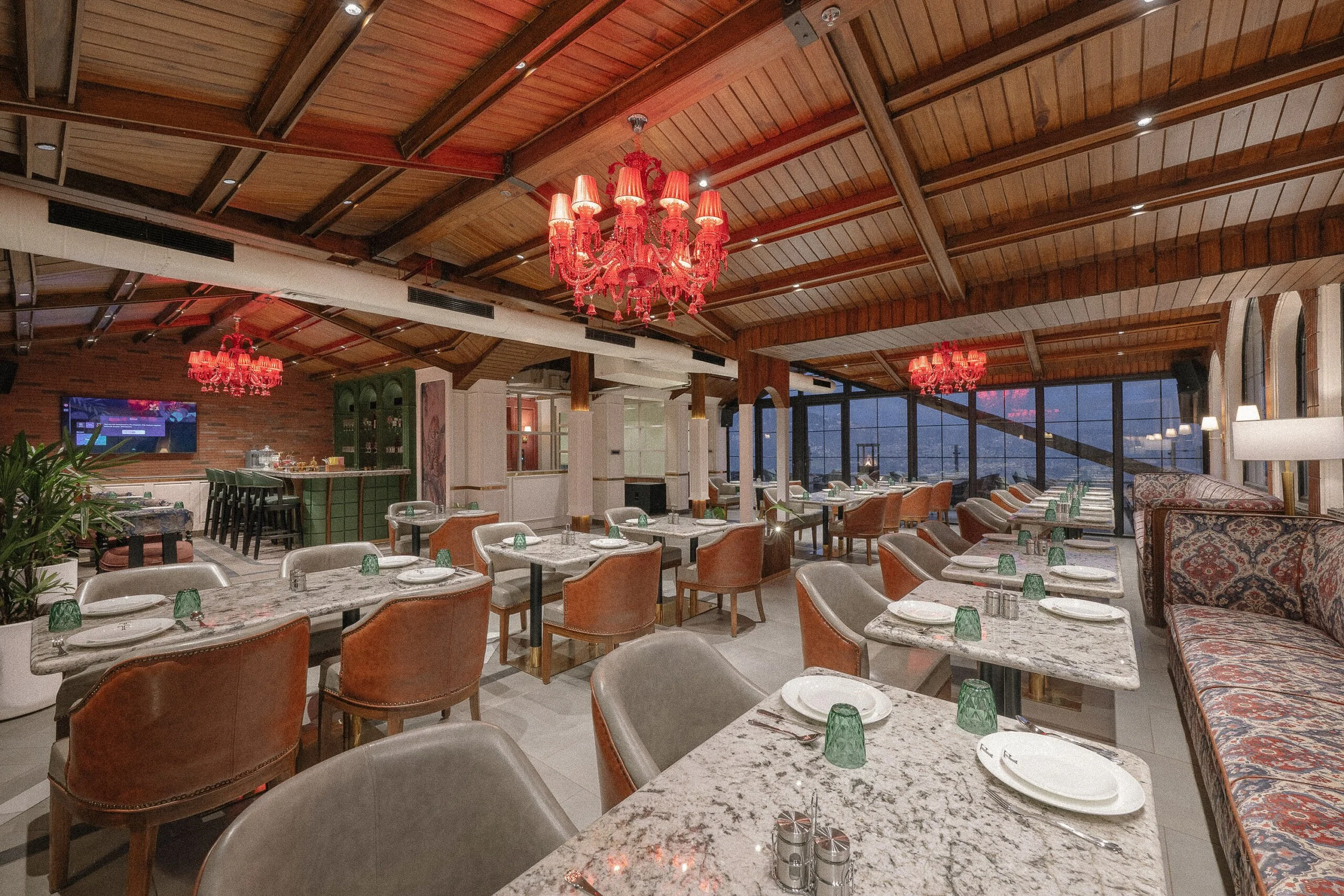 A spacious restaurant interior with a high wooden ceiling, red chandeliers, and large windows offering a city view. The dining area has marble-top tables set with plates, silverware, and green glass cups, surrounded by chairs and upholstered sofas.