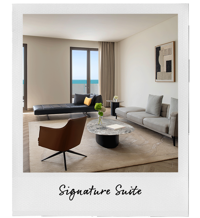 Modern hotel living room with a view of the ocean, featuring a black sofa, light gray sofa, brown chair, round marble coffee table with flowers, and a balcony door with curtains.
