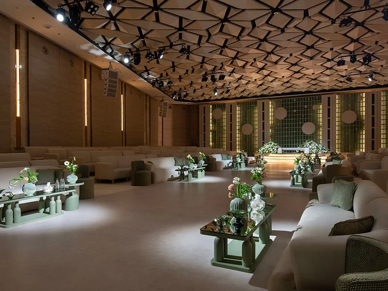 Empty, modern event space or lounge with beige sofas and green tables decorated with flowers and vases, ceiling with geometric patterns, stage at the front decorated with green curtains and white round lights, dim lighting.