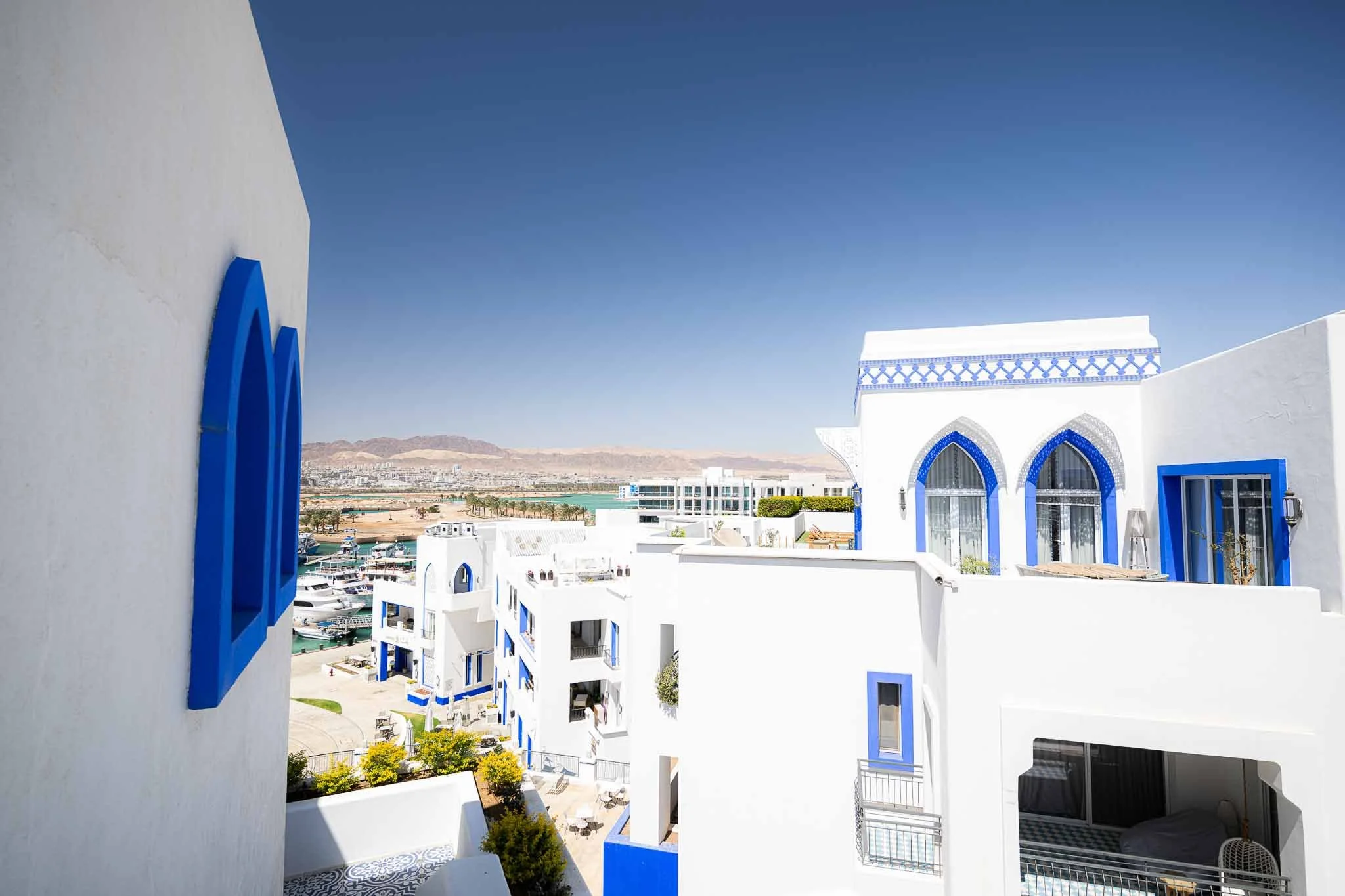 White buildings with blue accents and arched windows, overlooking a marina with boats and a distant mountain range under a clear blue sky.