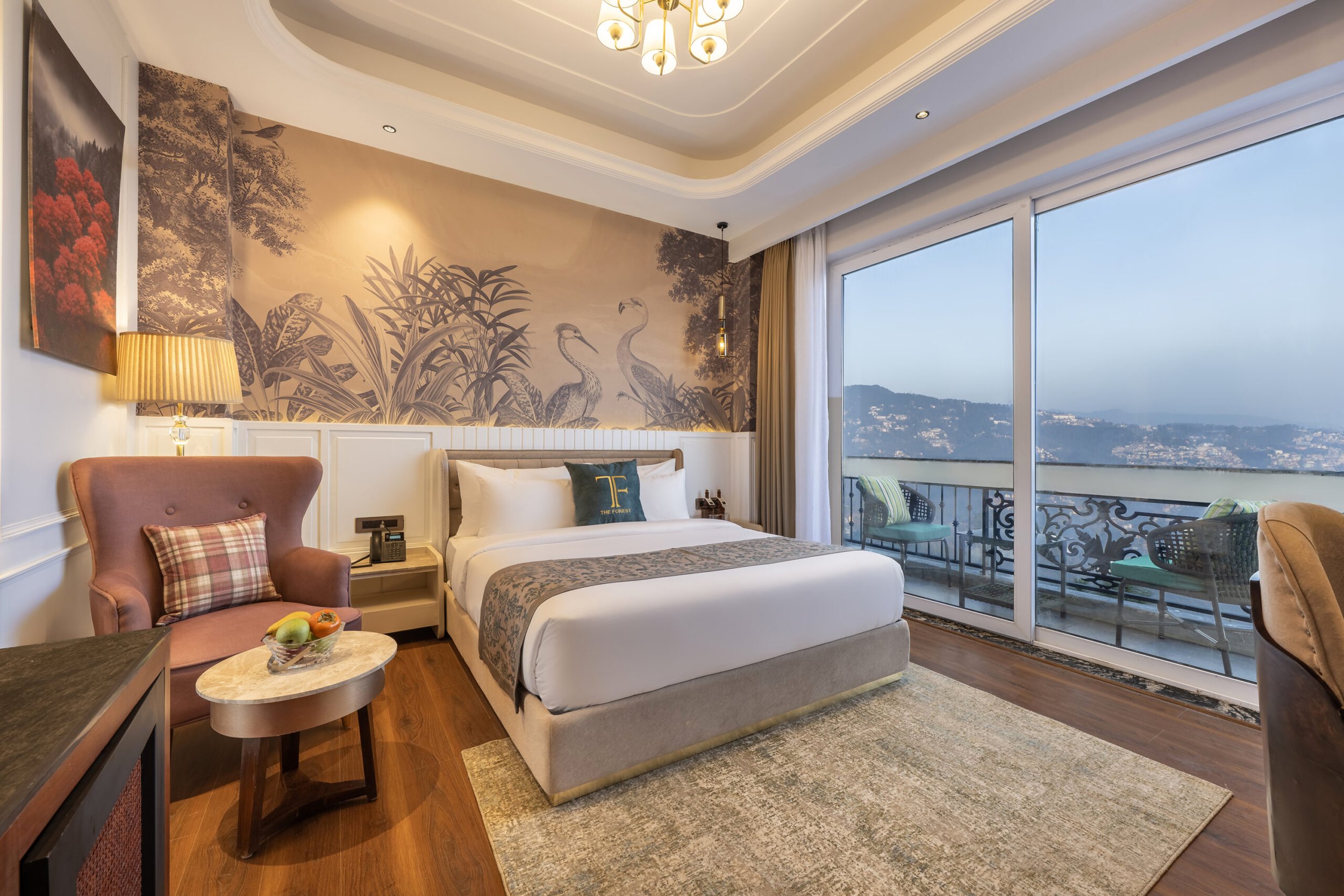 A hotel room with a bed, a pink armchair with a plaid pillow, a small round table with a fruit bowl, a wall mural of cranes and foliage, large window with balcony and city view, and warm lighting.