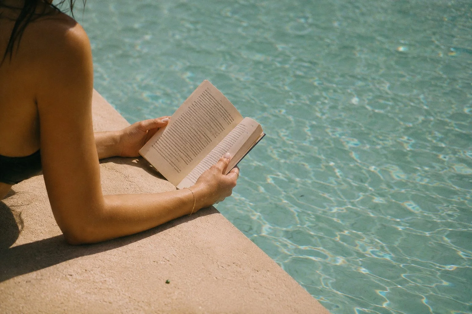 Person reading a book while lounging by a swimming pool.