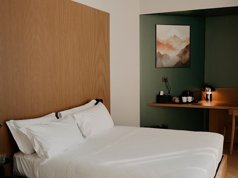 A hotel room with a bed, white bedding and pillows, a wooden headboard, a green accent wall with a landscape painting, and a small wooden desk with a lamp, coffee cups, and a black coffee maker.