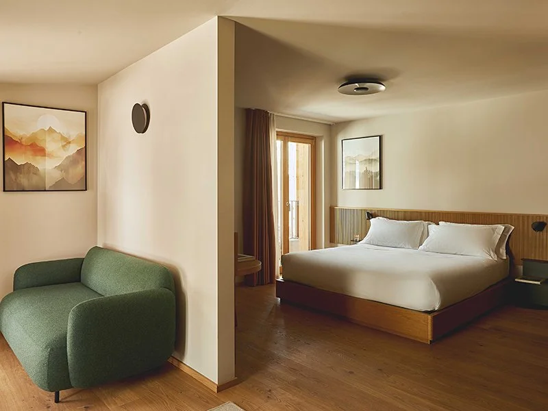 A cozy hotel room with a king-sized bed, wooden headboard, and a sitting area with a green sofa. The room has wooden floors, framed artwork on the walls, and a balcony door with curtains.