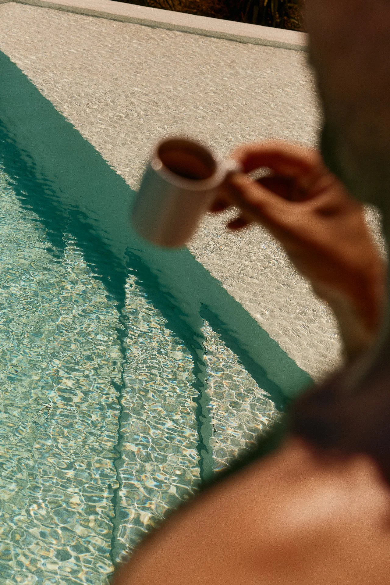A person holding a small plastic cup near a swimming pool. The photo is taken through a hole or opening, showing the pool and water ripples.