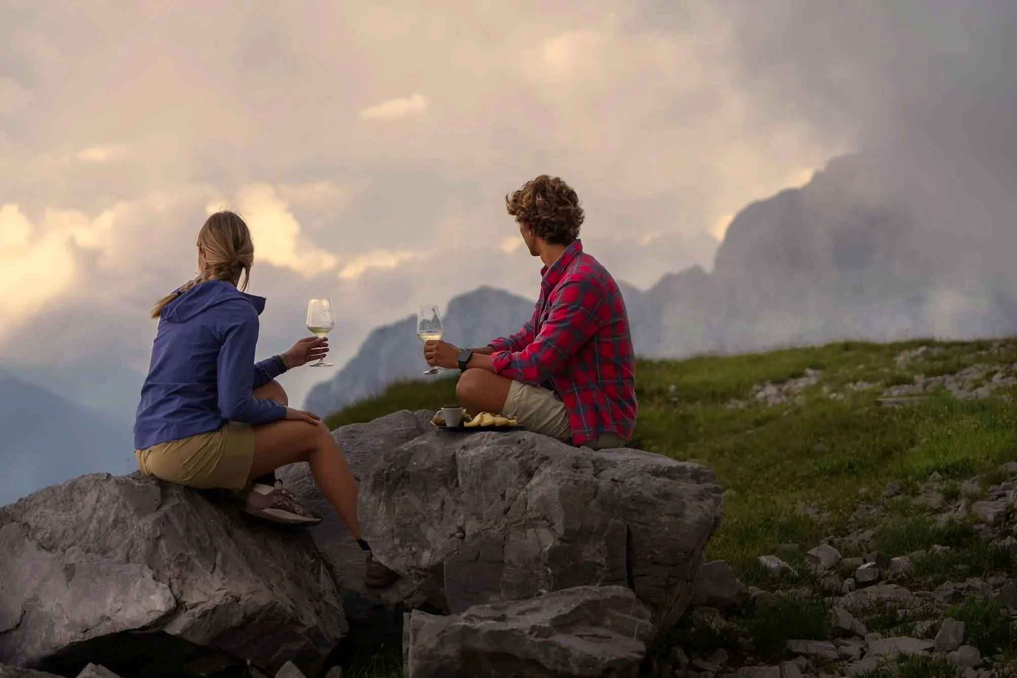 A man and a woman sitting on rocks outdoors, holding glasses of white wine, enjoying a scenic mountain view during sunset or dusk.
