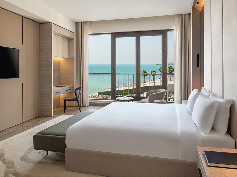 Beachside hotel room with a large window view of the ocean, balcony with chairs and palm trees, bed with white linens, small side table, and a sitting area.