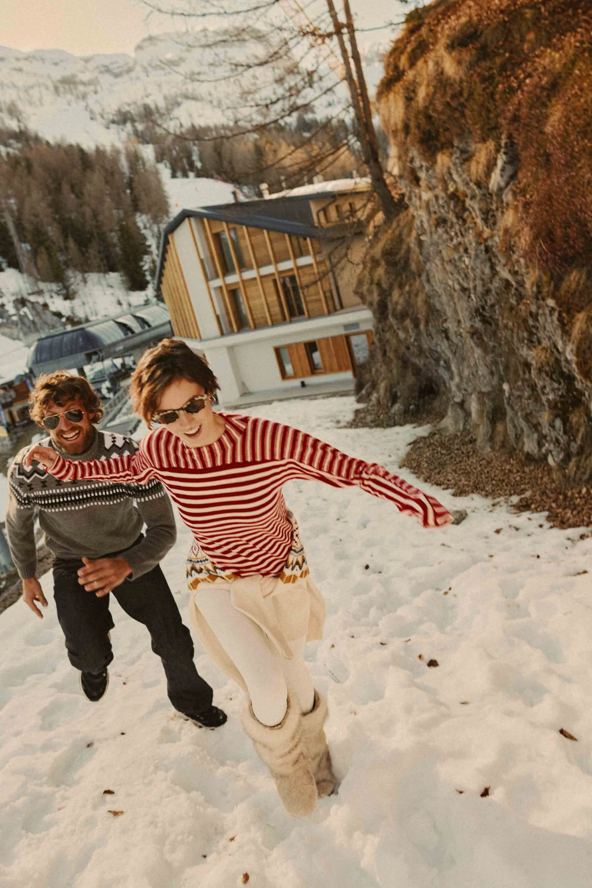 Two people, a man and a woman, smiling and running in snow outdoors with mountains and modern house in the background.