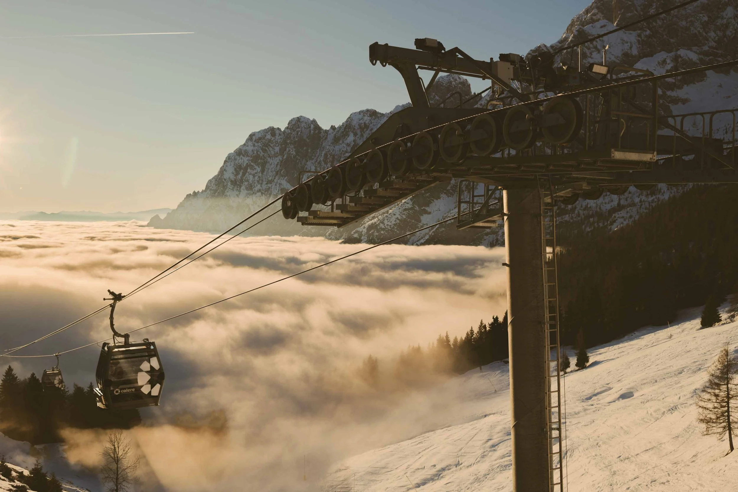 Ski lift chairs hanging from cables over snowy mountain landscape with clouds below and mountain peaks in the background at sunset.