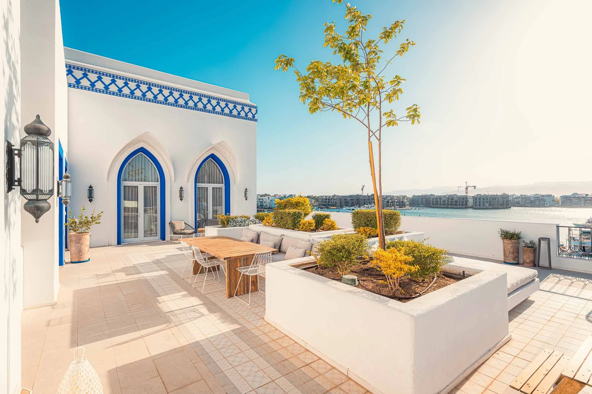Outdoor terrace with potted plants, dining table with chairs, white building with blue trim, and a view of water and modern buildings in the background.