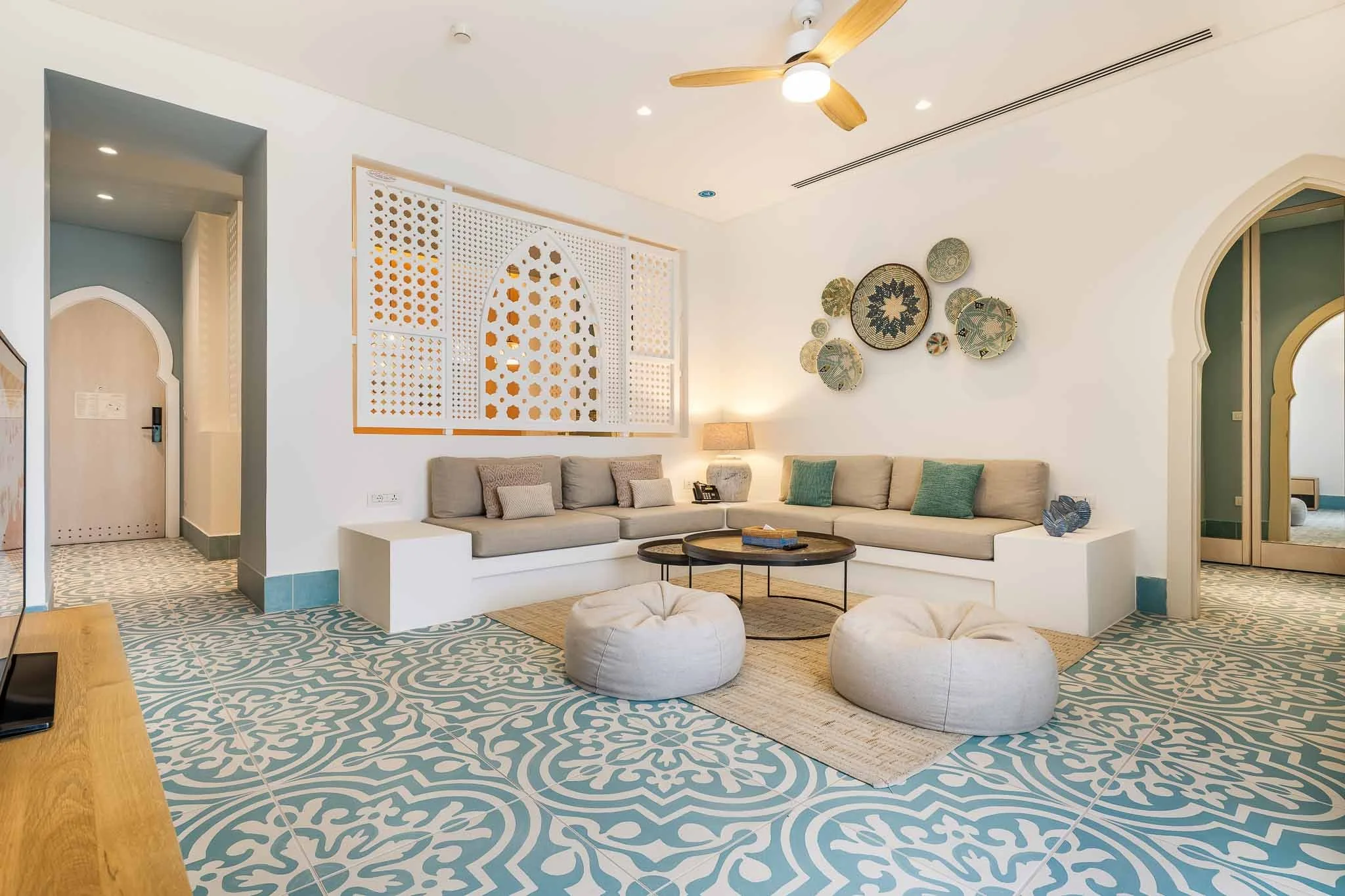 A cozy hotel lounge with a large L-shaped beige sofa, decorative pillows, a wooden coffee table, and two plush ottomans. The room features patterned blue and white tile flooring, a wall with decorative baskets, and soft lighting from table lamps and a ceiling fan with wooden blades.