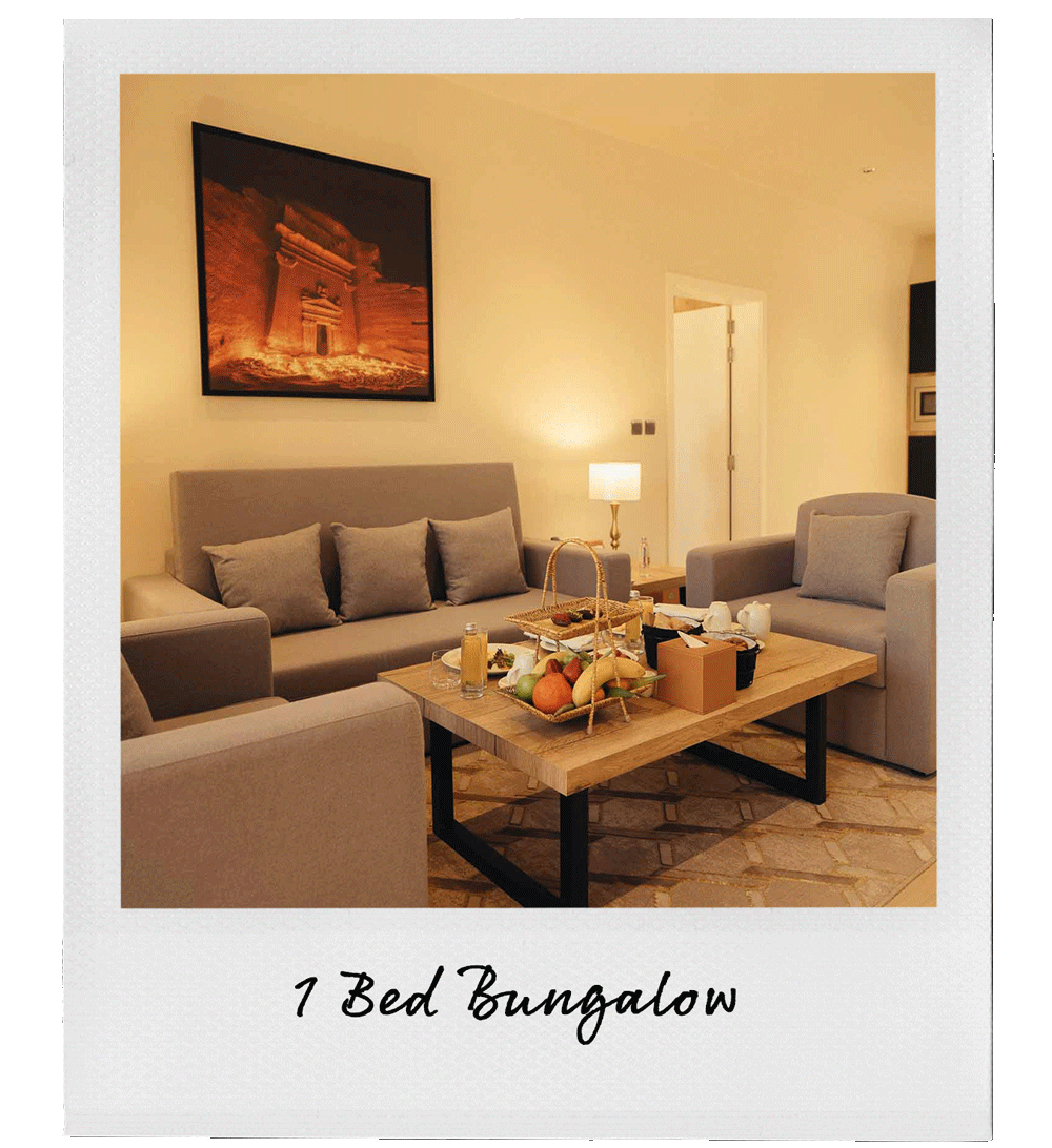 Living room with beige sofa, armchair, side table, and a wooden coffee table set with fruit, teapot, and snacks; artwork on wall; lamp providing warm lighting; white walls; part of doorway visible.