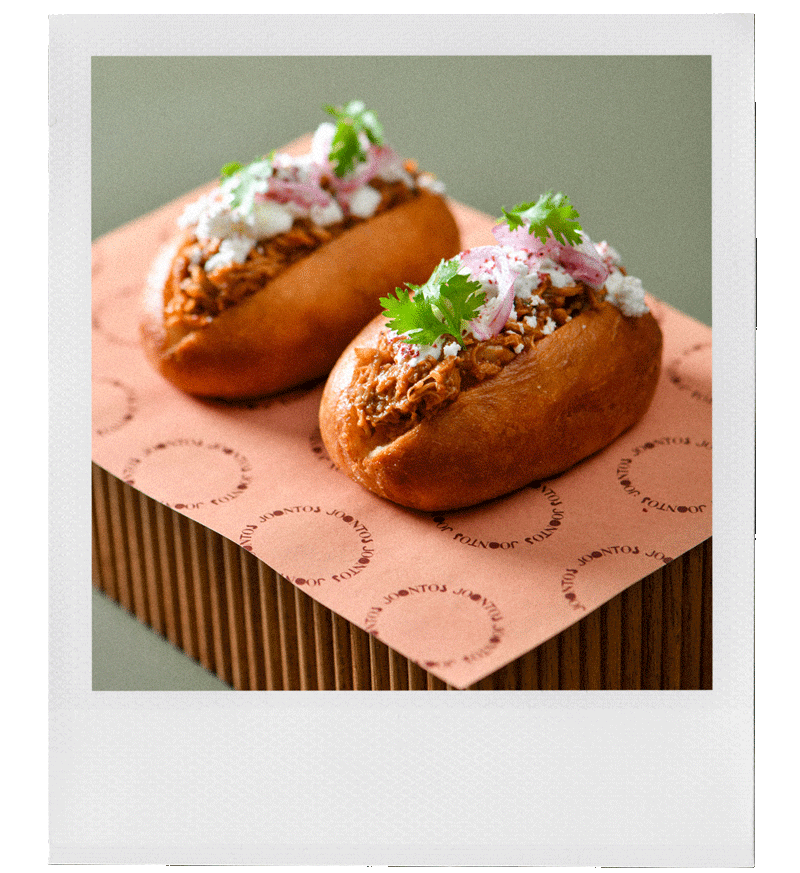 Due tacos con carne sfilacciata, conditi con salsa rosa e coriandolo, serviti su carta rosa con la scritta “JOUR” ripetuta più volte, adagiati su una superficie di legno.