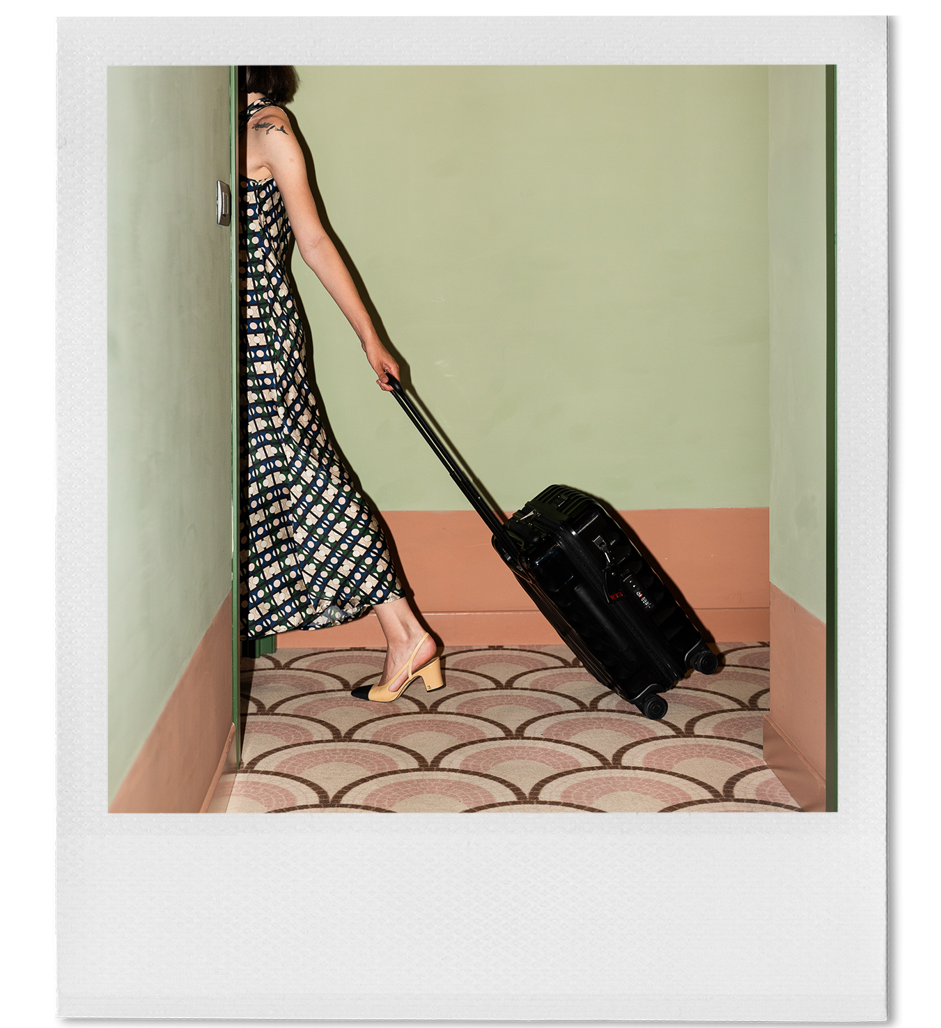 Woman in a patterned dress and heels pulling a rolling black suitcase into a room through a doorway.