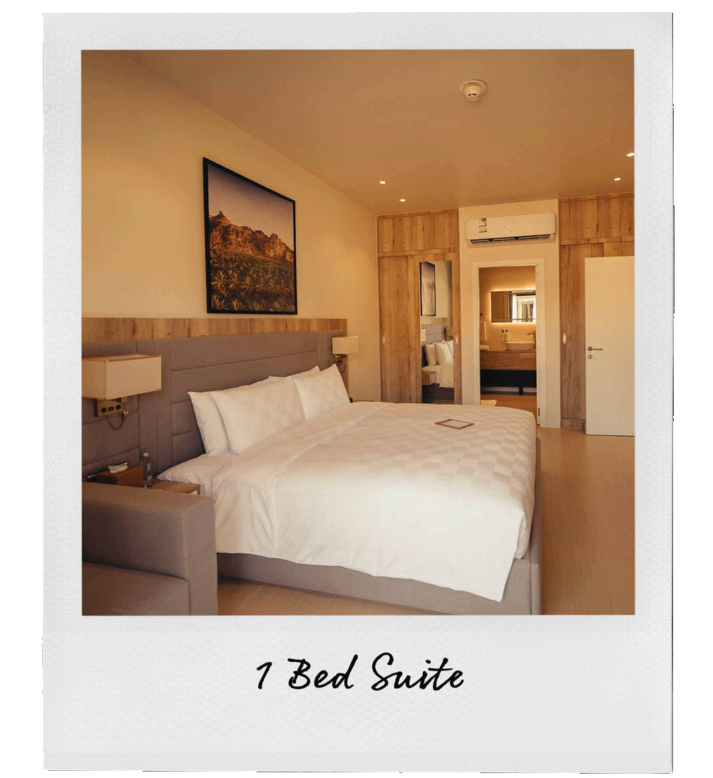A modern hotel bedroom with a large bed, wall-mounted lamps on either side, and a framed landscape painting above the bed. There is a small tablet on the bed and an air conditioning unit above the doorway that leads to a bathroom. The room has light wood accents and neutral-colored walls.