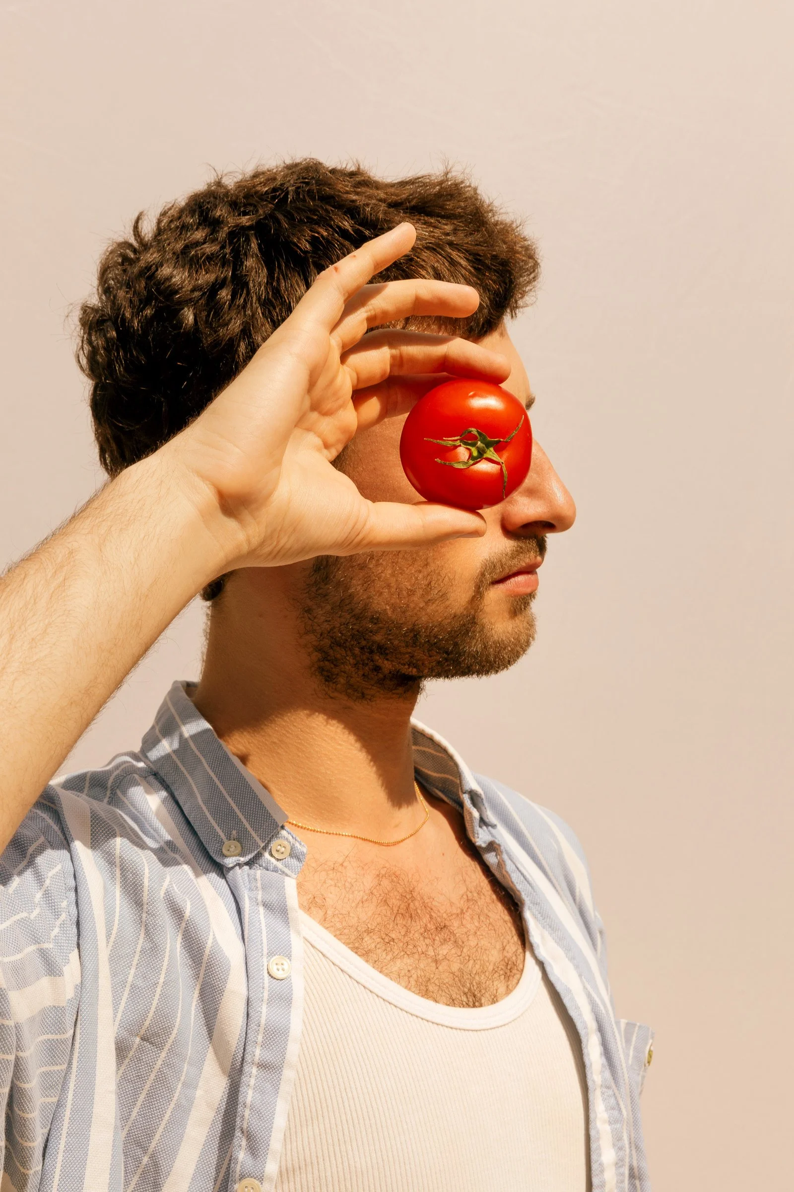 A man with dark curly hair and light skin holds a red tomato in front of his face, covering one eye and part of his nose, against a plain light background. He is wearing a light-colored striped shirt over a white tank top.