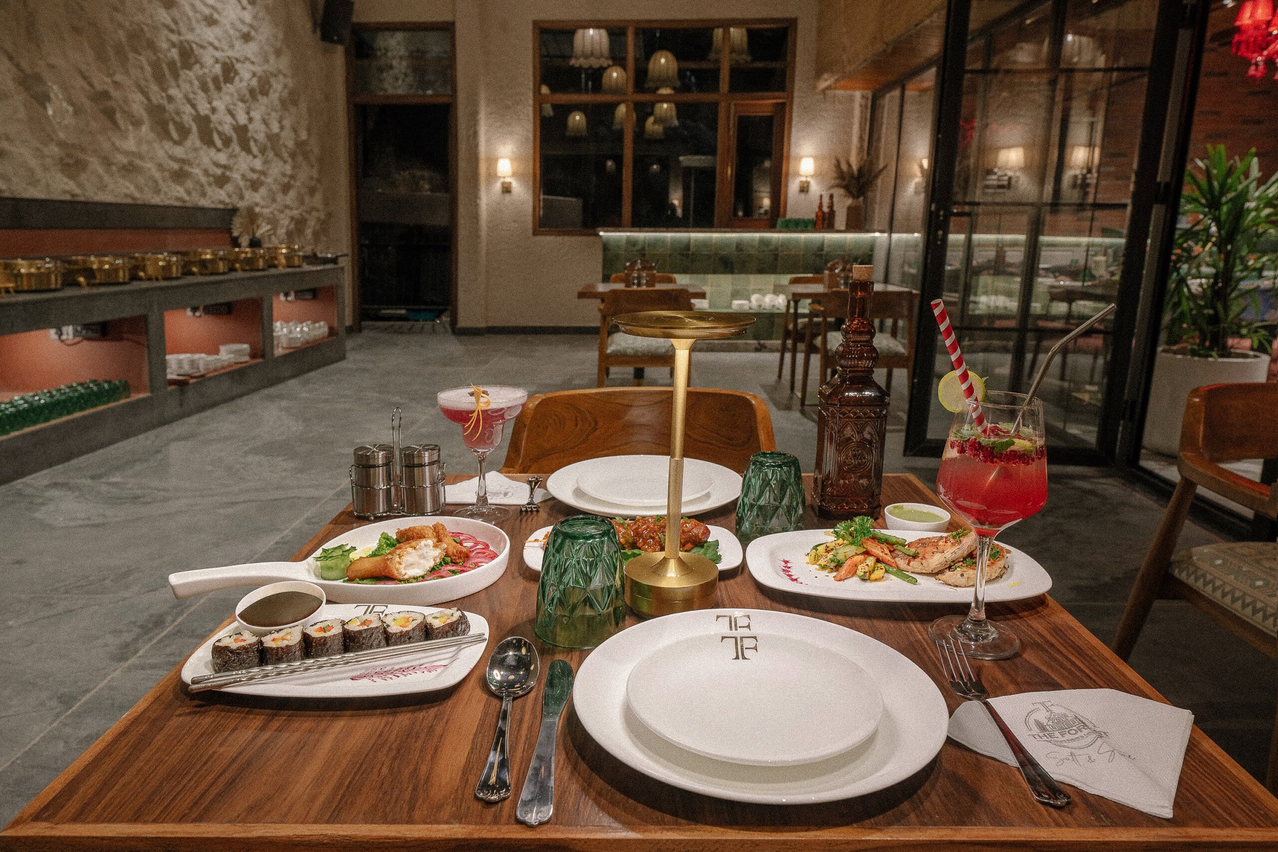 A table set with various Asian dishes including sushi rolls, spring rolls, and grilled chicken, in a restaurant with ambient lighting, decorated with green glassware and a drink with a striped straw.