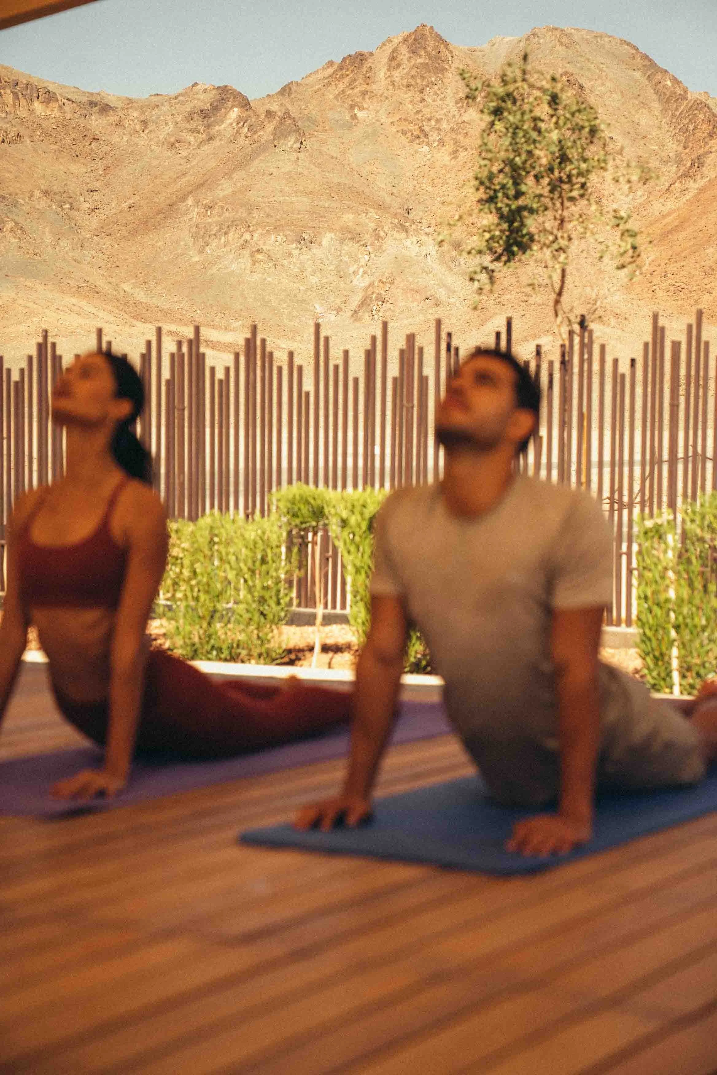 Two people practicing yoga outdoors on their mats, with a desert mountain range in the background.