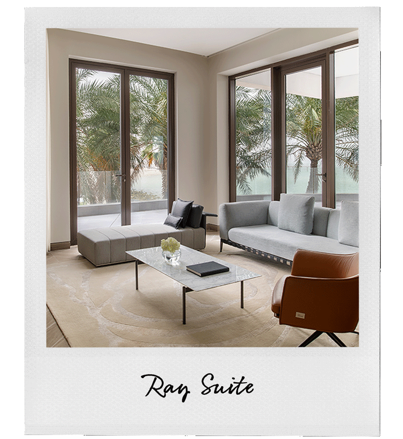 Modern living room with large glass sliding doors and windows, overlooking palm trees, containing a gray sofa, a tan armchair, a marble coffee table with a flower vase and book, a beige area rug, and throw pillows.