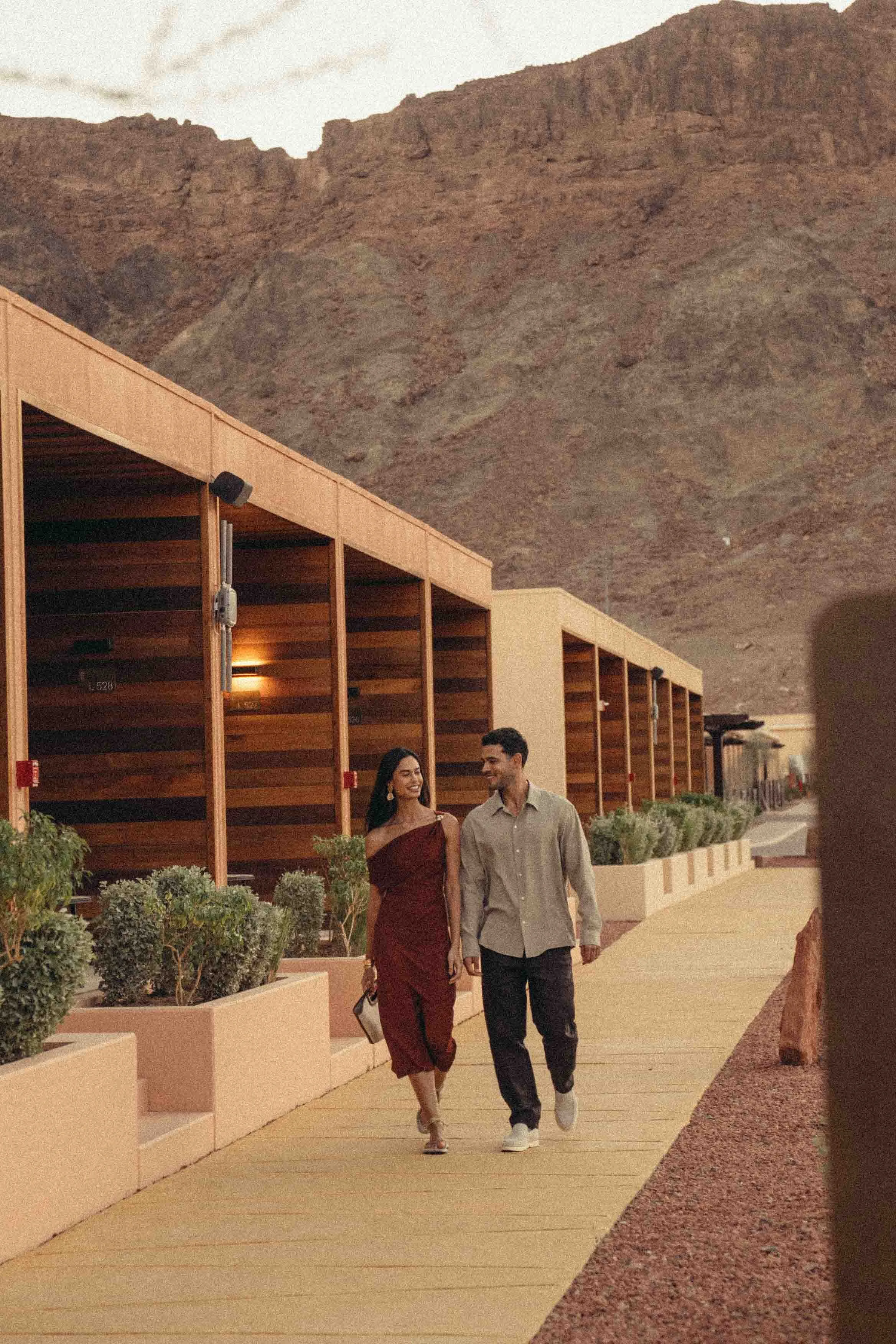 A man and a woman walk hand in hand outdoors in front of a mountain backdrop, with modern architectural structures and landscaped surroundings.