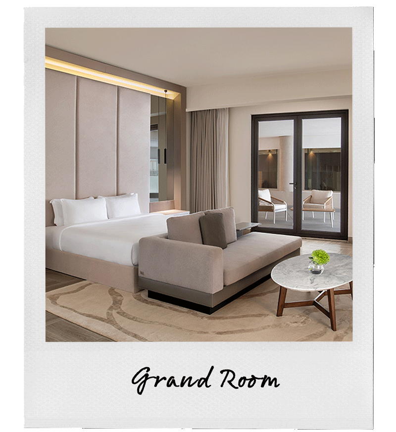 A luxurious hotel Grand Room with a large bed, a gray sofa facing the bed, a round marble coffee table with a green plant, and a balcony with chairs visible through glass doors.