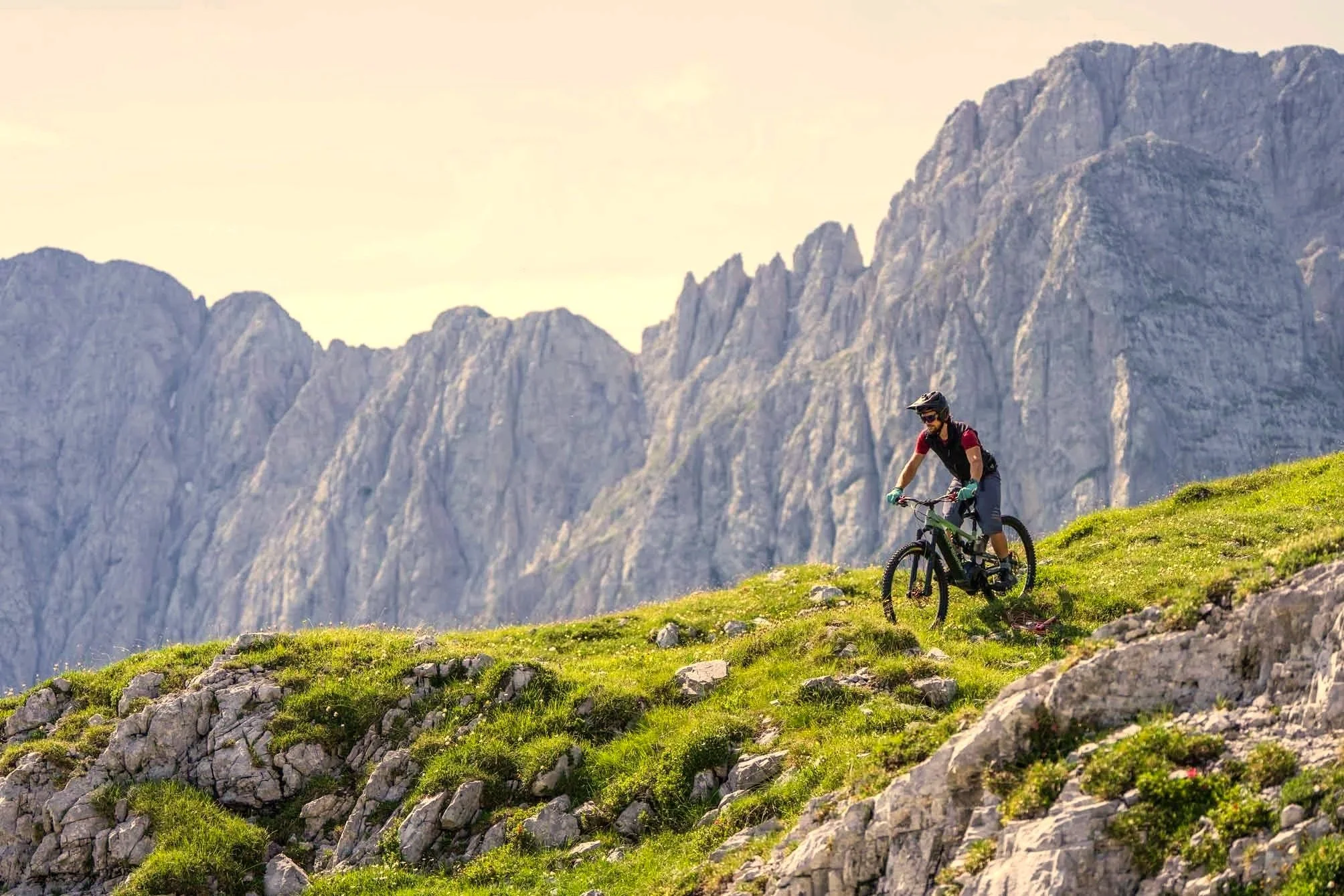 A person mountain biking on a green grassy slope with rocky terrain and mountains in the background.