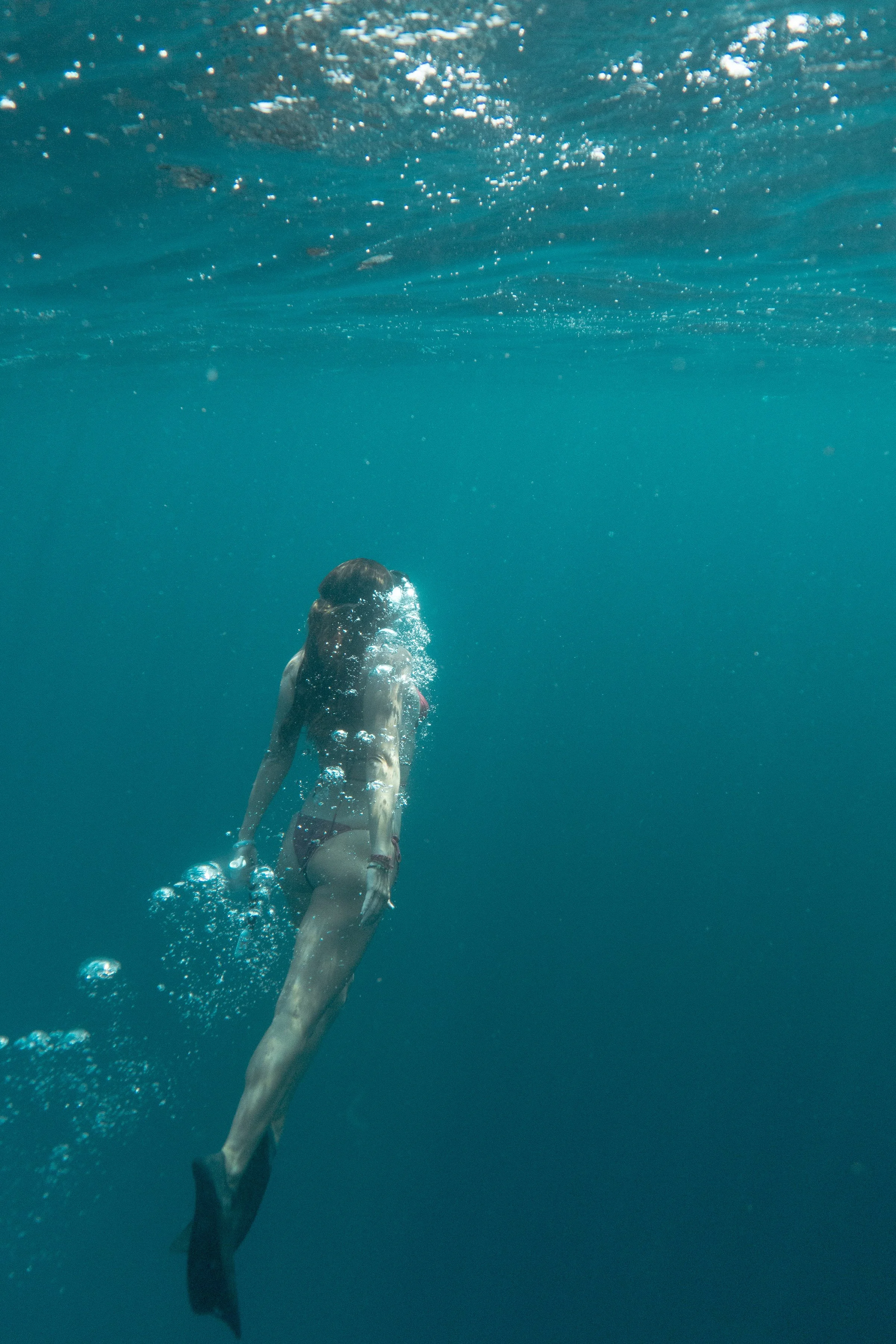 A person swimming underwater, seen from below, with their back and legs visible and bubbles emerging around them.