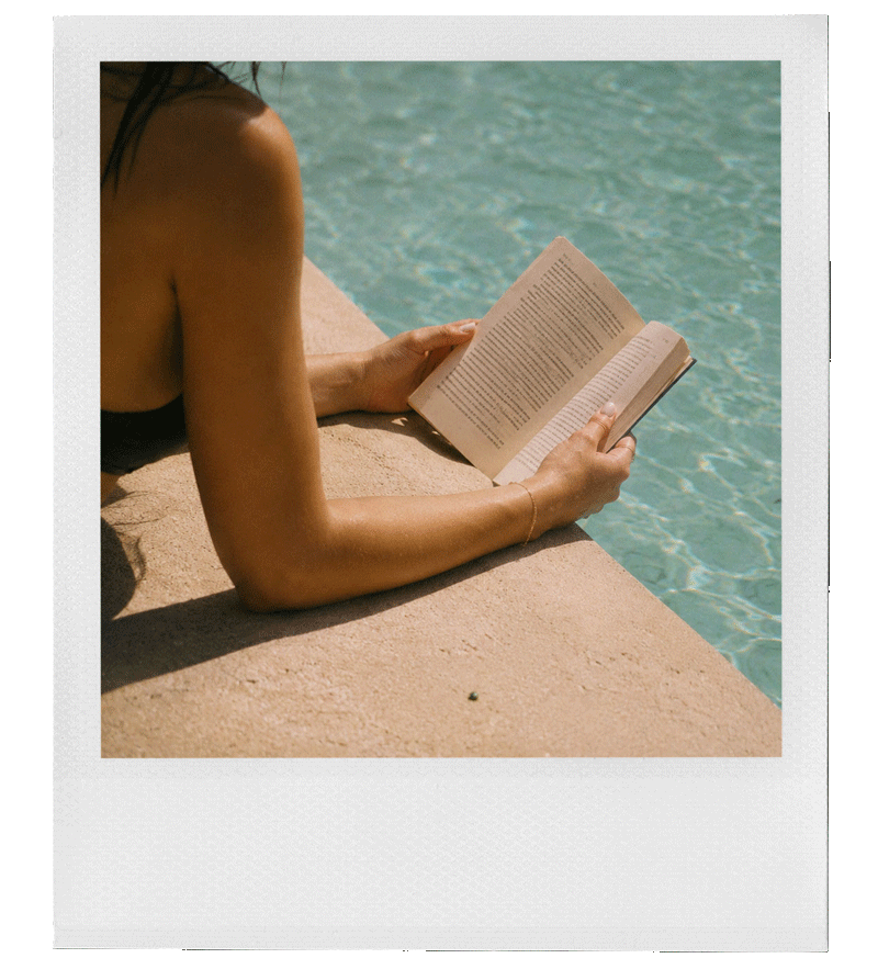 A woman is lying on her stomach on the edge of a pool, reading a book, with the water visible in the background.