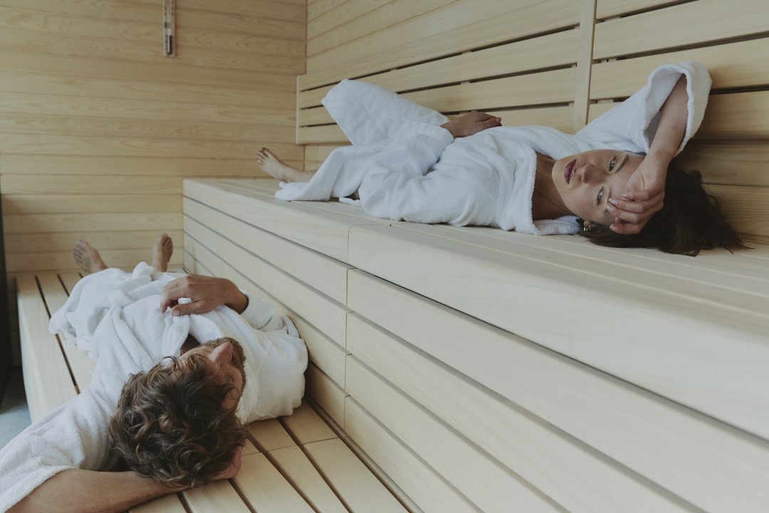 Two women in robes lying on benches inside a wooden sauna, relaxed and resting.