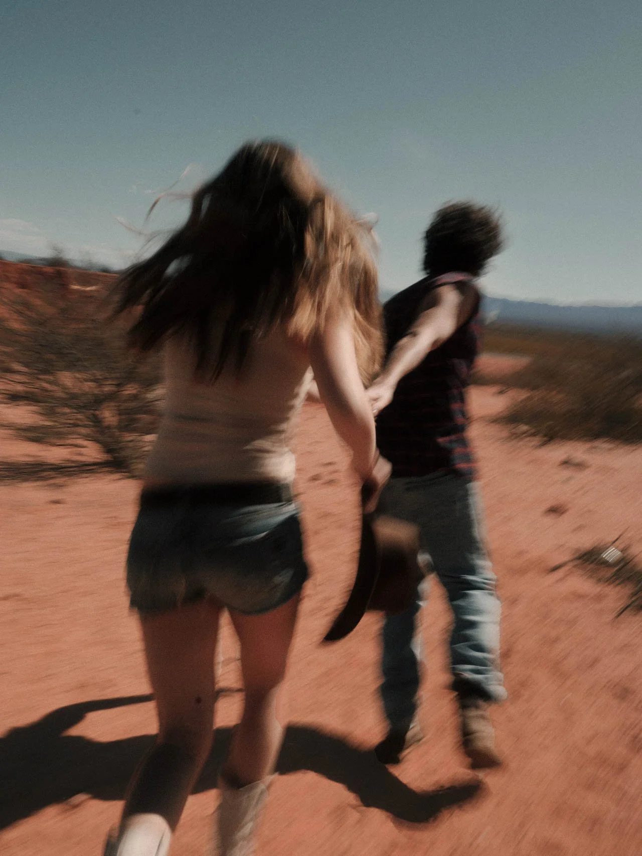 A young woman with long hair and a young man hold hands as they walk on a dirt path in a desert landscape during the daytime.
