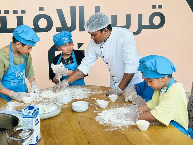 Chef instructing children on baking, kids wearing blue chef hats and aprons, working with flour on a wooden table.