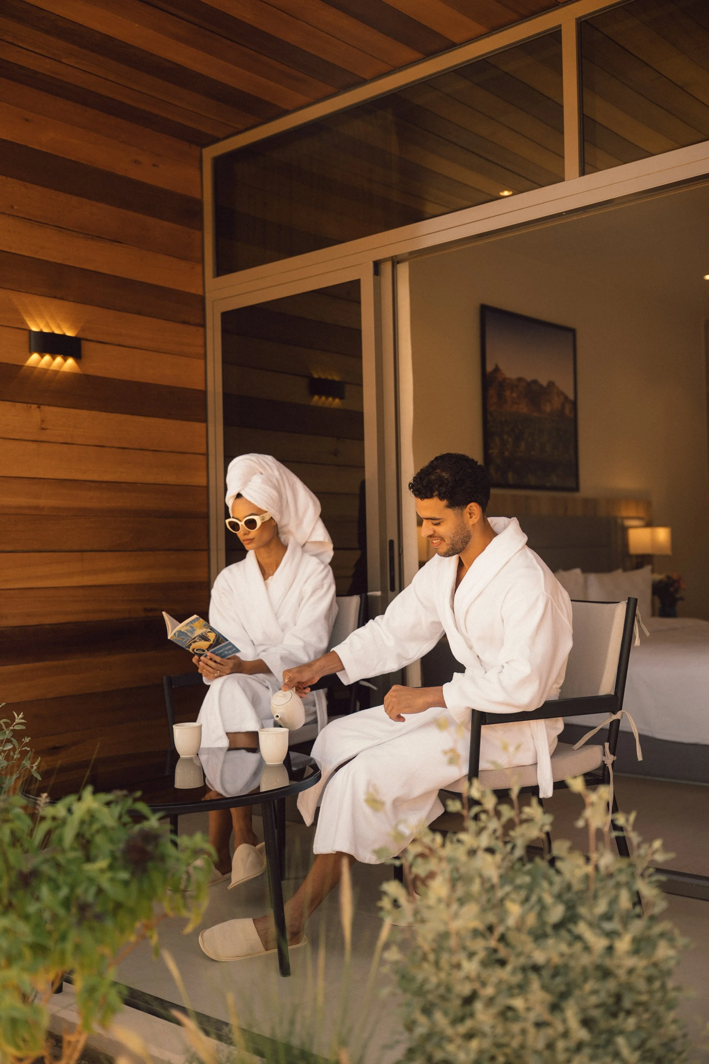 Two people, a man and a woman, sitting on a balcony or patio, dressed in white bathrobes, with the woman wearing sunglasses and a towel wrapped around her head, and the man pouring a beverage into a cup. There are cups on the table and some greenery in the foreground, with warm lighting and wooden paneling on the wall.