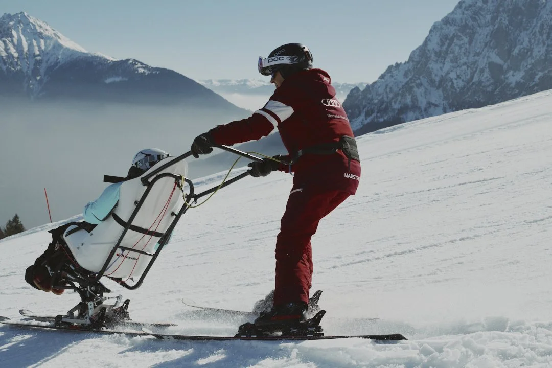 Person in red ski gear pushes a sled with a child down snowy mountain slope in a mountain landscape with snow-covered peaks.