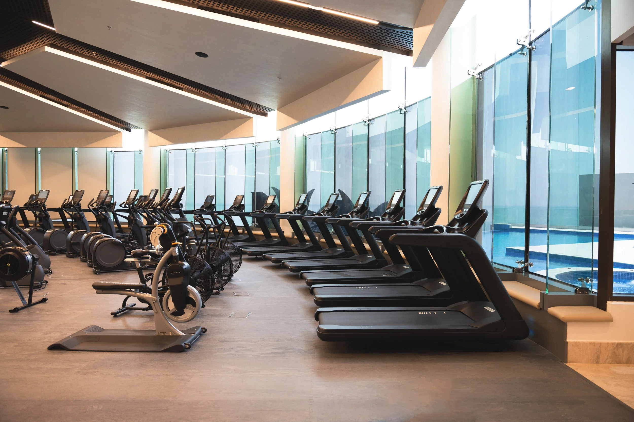 Rows of treadmills and exercise bikes in a modern gym with large windows and a pool visible outside.