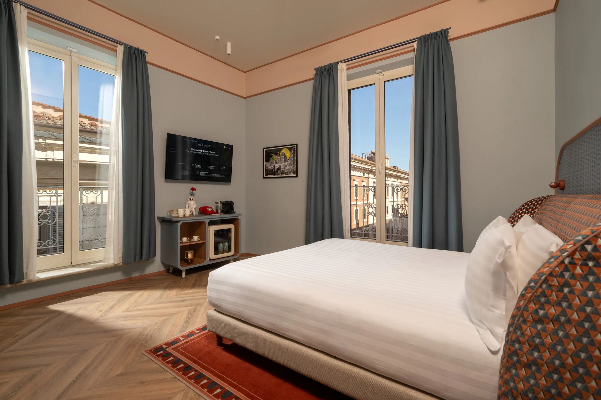 A hotel room with a large bed, a flat-screen TV on the wall, and a glass door with curtains leading to a balcony. The room has light-colored walls, wooden trim, a small table with cups and a coffee maker, and framed pictures on the wall. The balcony shows neighboring buildings with tiled roofs.