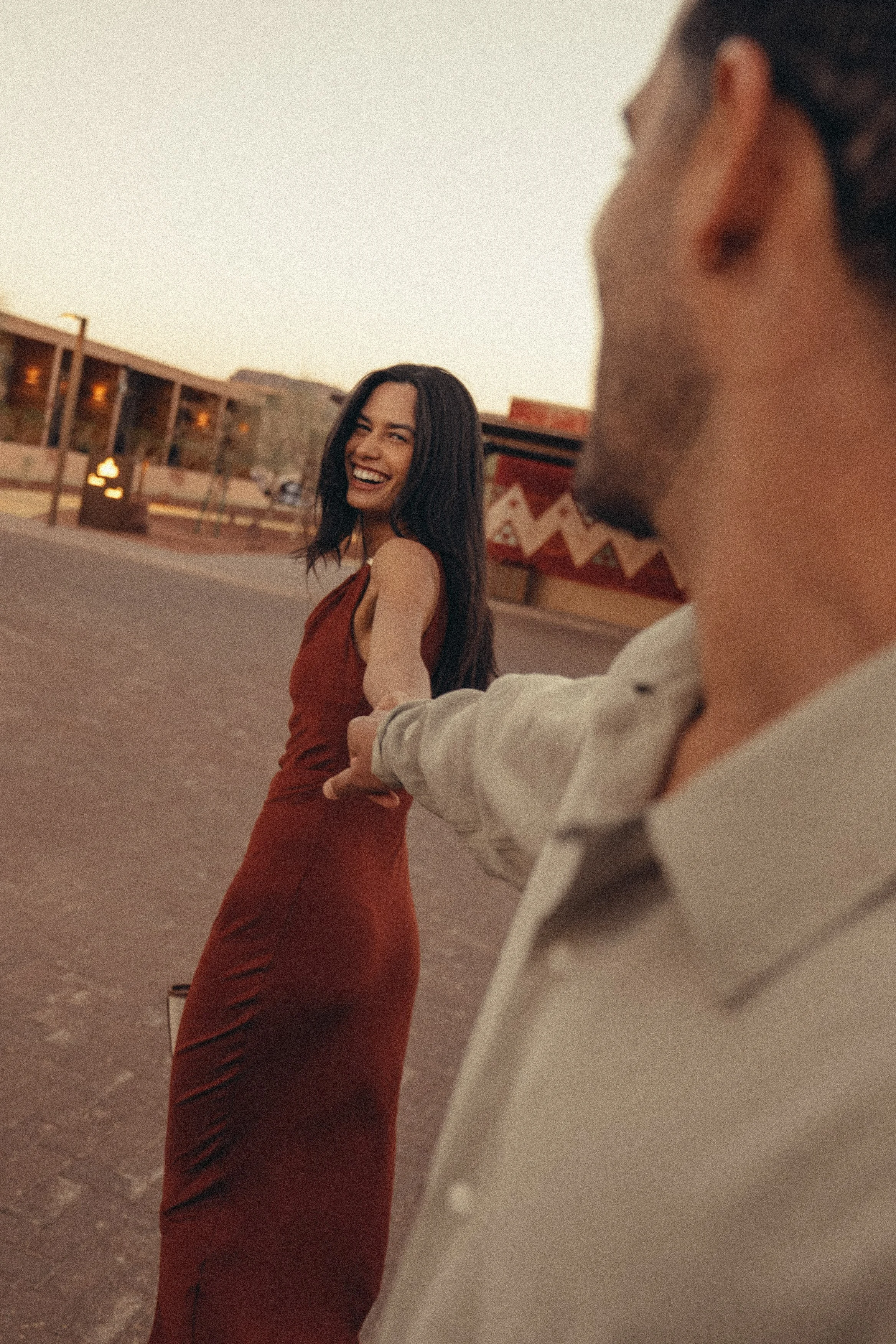 A woman in a red dress holding hands with a person in a white shirt, smiling and looking back at the camera with a sunset in the background.