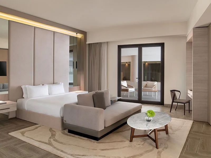 Modern hotel room with a large bed, a sofa, a coffee table with a green plant, a chair, and a balcony with outdoor seating.