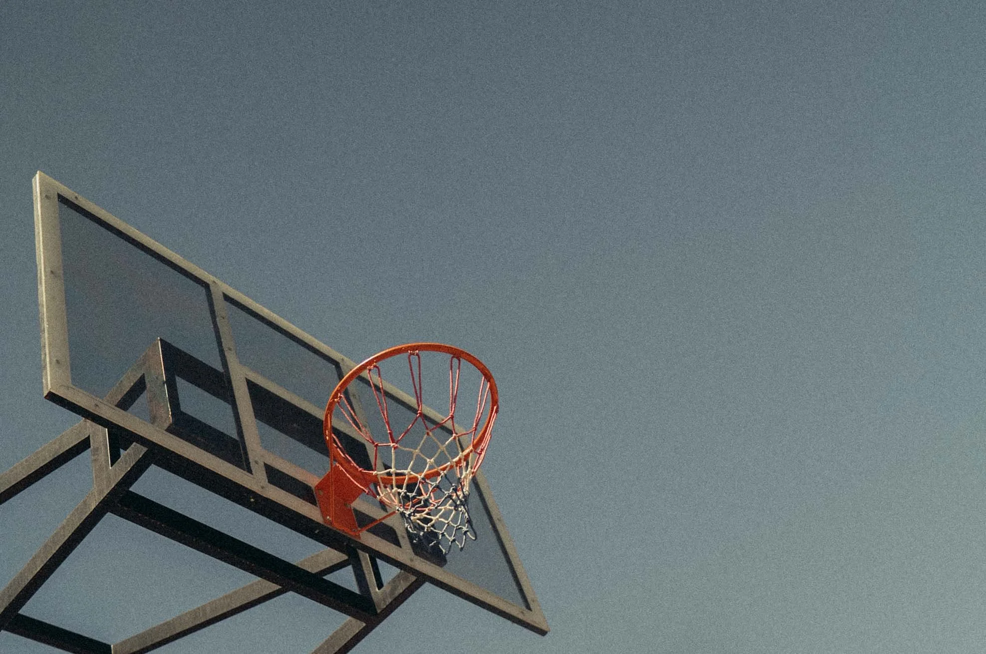 A basketball hoop with an orange rim and net, mounted on a metal backboard structure, against a pale blue sky.