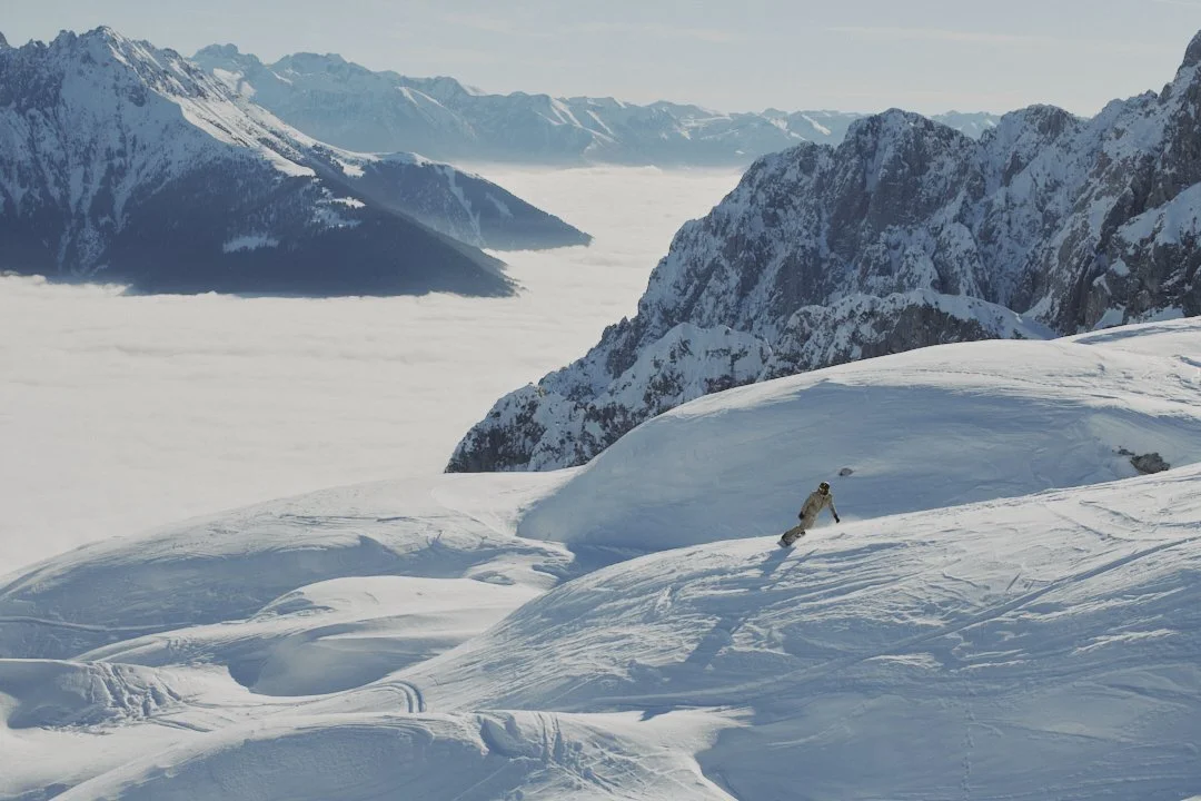 A person skiing on a snow-covered mountain slope with mountain peaks in the background and clouds below.
