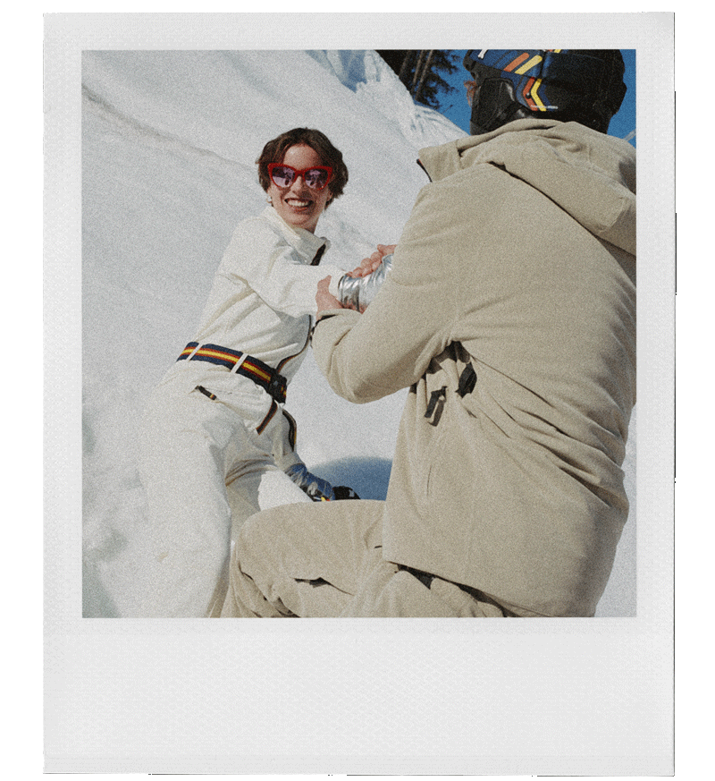 A woman with short brown hair and red glasses smiling while holding hands with a man in a beige ski jacket on a snowy mountain slope.
