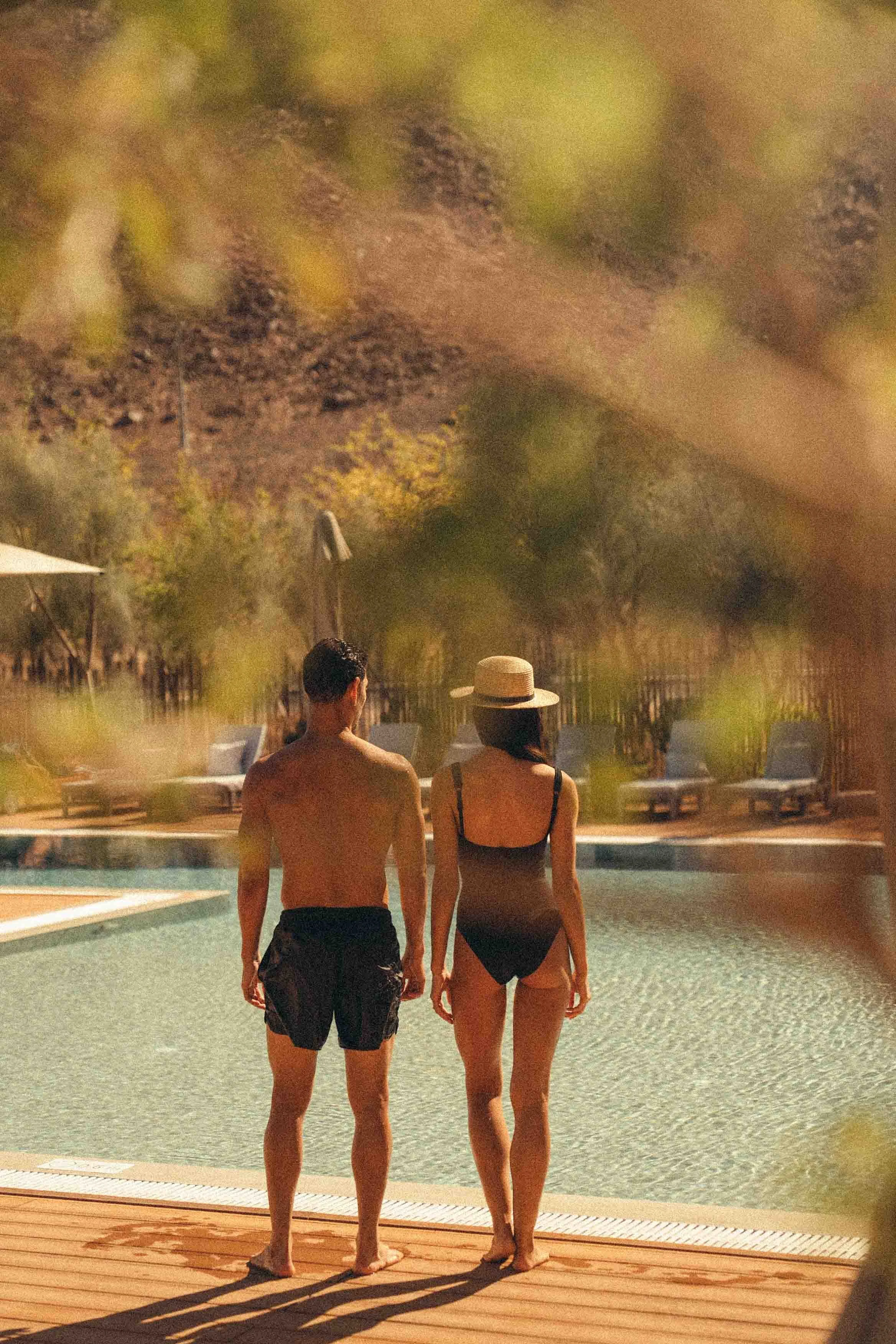 A man and woman in swimwear standing by a swimming pool during evening time, with lounge chairs and umbrellas in the background. They are holding hands and facing the pool.