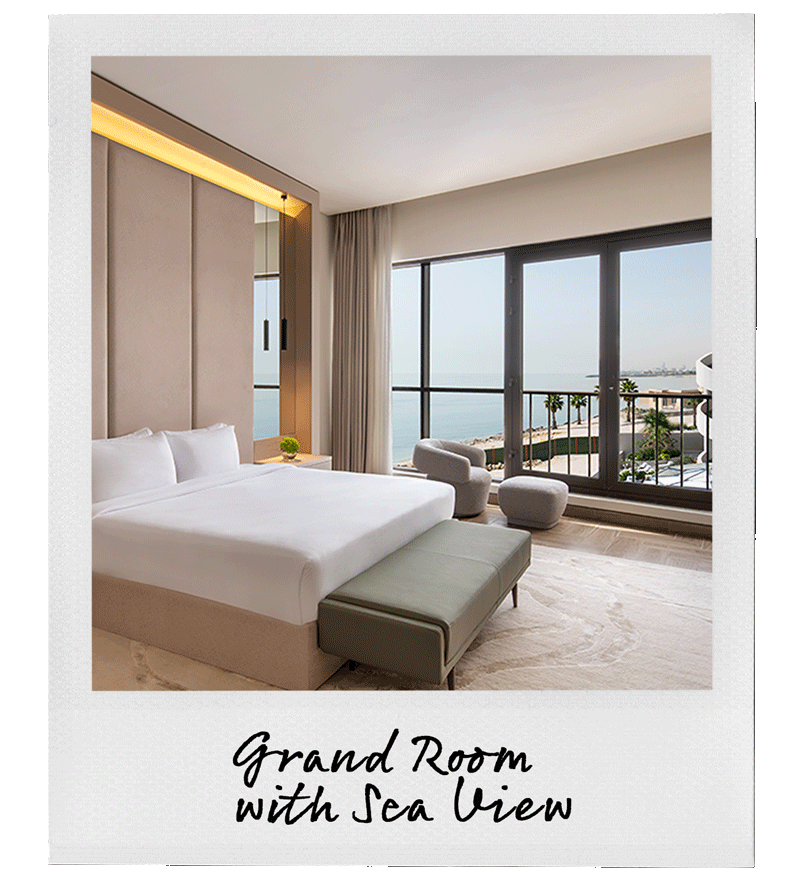 Modern hotel room with a large bed, a bench at the foot, and two chairs near floor-to-ceiling windows with a sea view.