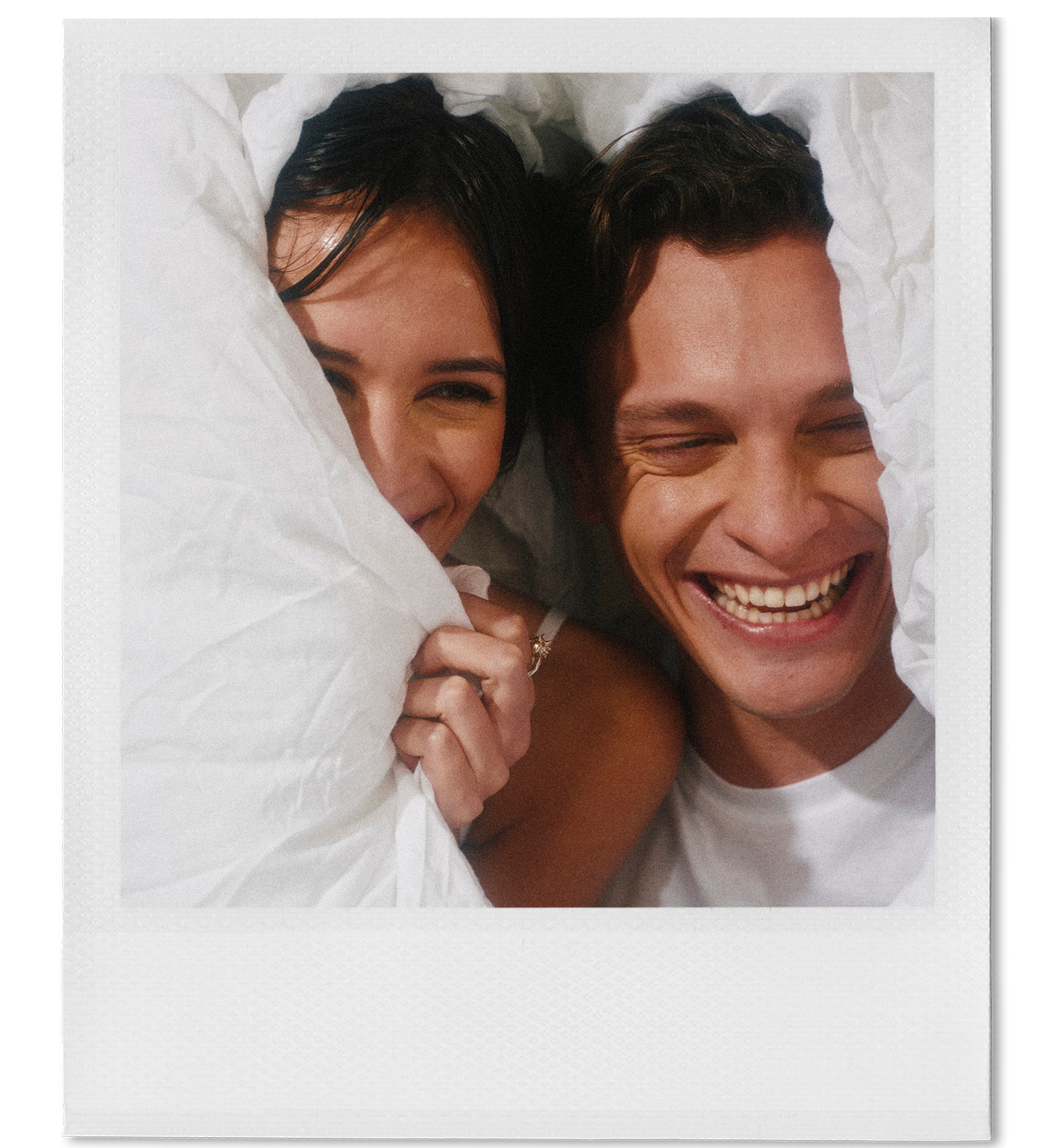 Two people smiling and laughing under white sheets in bed, appearing to enjoy a joyful moment together.