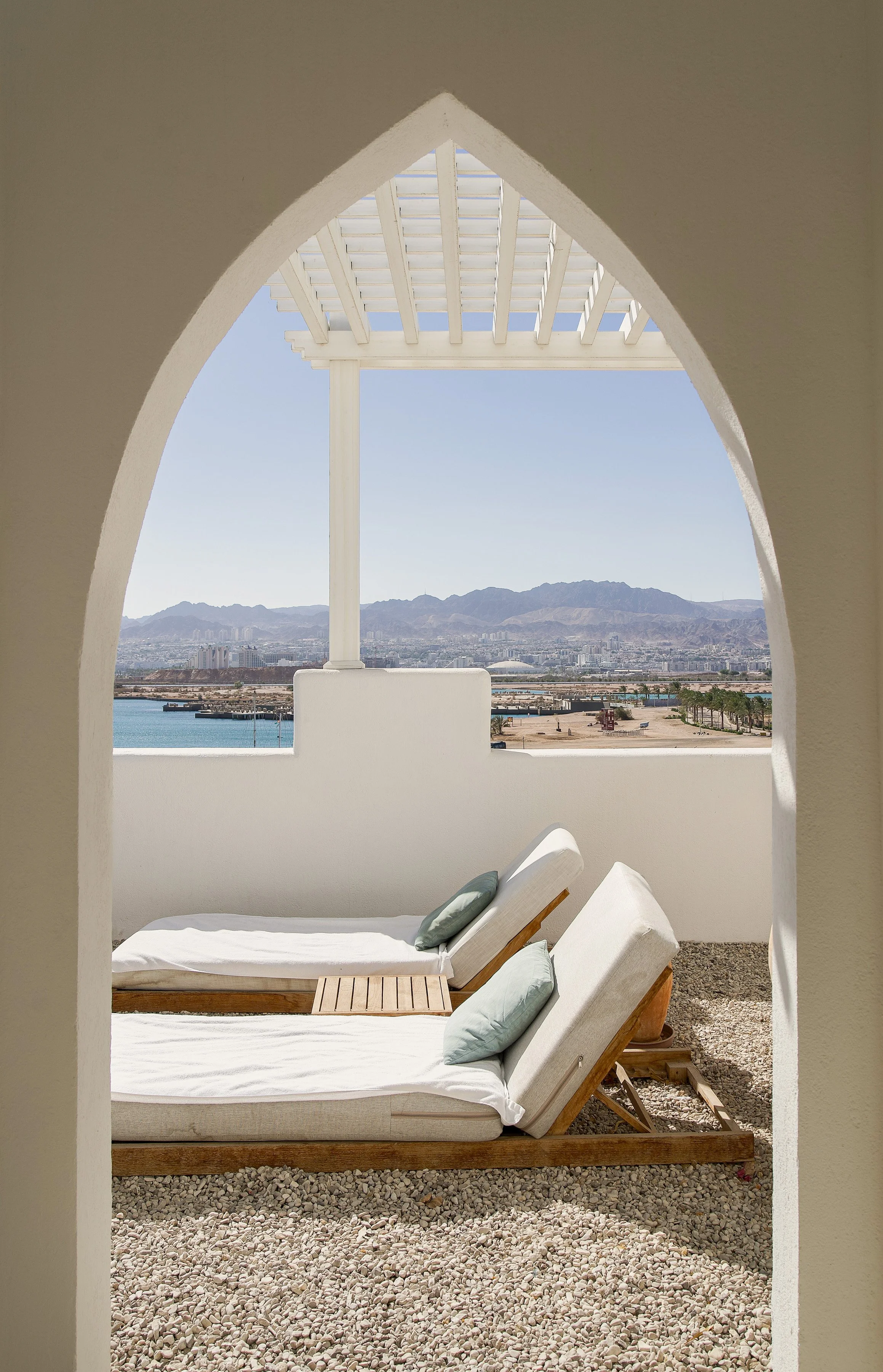Outdoor seating area with white lounge chairs and cushions, overlooking a body of water and mountain range in the distance, with a white pergola overhead, framed by an arched architectural opening.