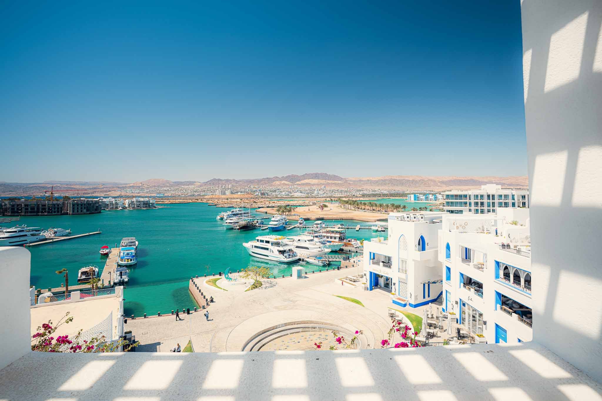 View of a marina with yachts and boats, white buildings with blue accents, cave-style architecture, and a clear blue sky, likely in a coastal resort area.