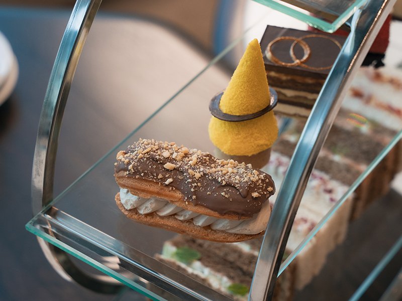 Assorted desserts displayed on a glass stand, including a chocolate eclair with crushed nuts, a yellow cone-shaped pastry, and layers of cake in the background.