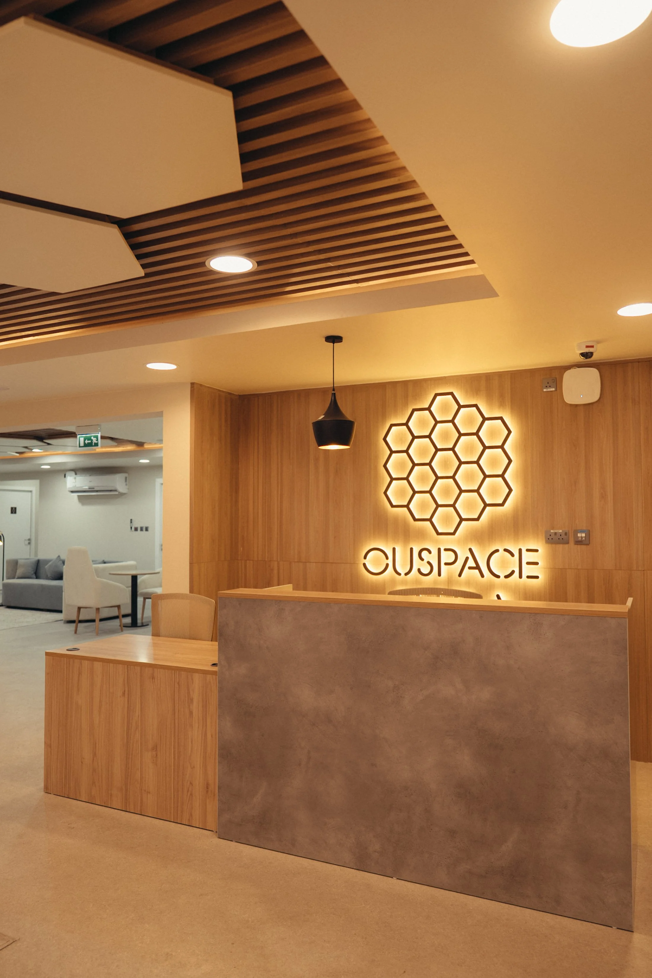 Interior view of a modern reception area with a wooden wall and backlit logo reading 'CUSPAC'. The ceiling has wooden slats, and there is a black hanging light fixture. In the background, there is a seating area with chairs and tables.