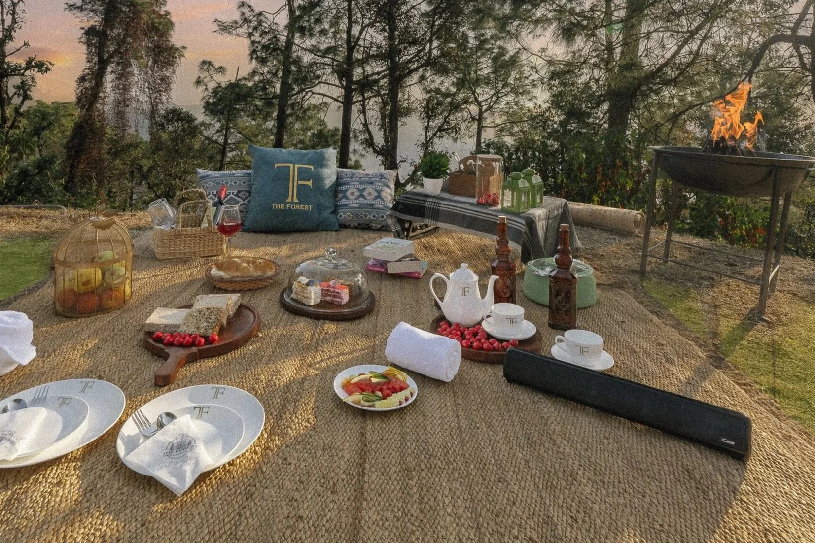 A picnic setup on a woven mat outdoors with plates, cups, a teapot, cake, fruit, and decorative pillows, with a fire pit in the background among trees.